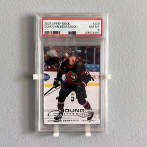 May include: A 2025 Upper Deck Donovan Sebrango ice hockey card, graded NM-MT 8 by PSA, encased in a clear plastic protective slab. The card features a player in a black Ottawa Senators jersey, with the text Young Guns and the player's name visible.