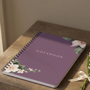 May include: A purple spiral-bound notebook with the word NOTEBOOK printed in white on the cover. The cover features floral illustrations in the corners with white and pink flowers and green leaves. A small bouquet of lily of the valley is tied with a white ribbon.