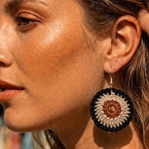 Peut inclure: Une boucle d'oreille crochetée circulaire présentant un motif concentrique avec un centre marron foncé, un anneau central beige clair et une bordure extérieure noire. La boucle est fixée à un crochet en métal argenté et pend au lobe.