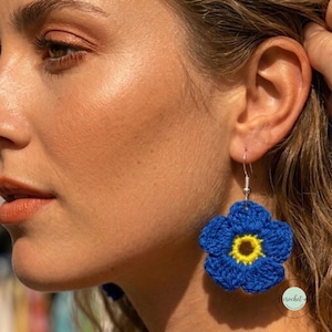 May include: A crochet flower earring featuring five rounded blue petals and a bright yellow center. The floral design is crafted from yarn and hangs from a silver-toned metal hook. The earring is shown worn on an earlobe, displaying the texture.