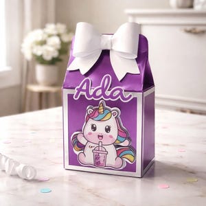 May include: A purple gift box with a white bow on top. The box features a cartoon unicorn illustration and the name "Ada" in script. The unicorn has a rainbow mane and is holding a drink.