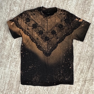May include: Black t-shirt with a bleached design. The shirt features a lace-like pattern across the chest and shoulders. The design is a mix of black and tan colors, with a speckled effect.