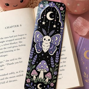 May include: A black bookmark with a whimsical design featuring a moth, mushrooms, stars, and crescent moons. The bookmark has a purple and white color scheme with floral accents. The text "CHAPTER 8" is visible on the open book.