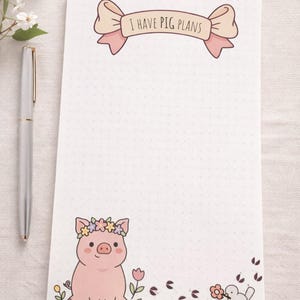 May include: A white notepad with a dotted pattern, featuring a cartoon pig wearing a flower crown. The notepad has the text "I HAVE PIG PLANS" above the dotted area. A silver pen rests on the left side of the notepad.