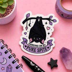 May include: A sticker featuring a black bat hanging upside down from a branch, with the text "Hanging on by Pure Spite" on a purple banner. The design includes hearts, stars, and a crystal, set against a pink background.