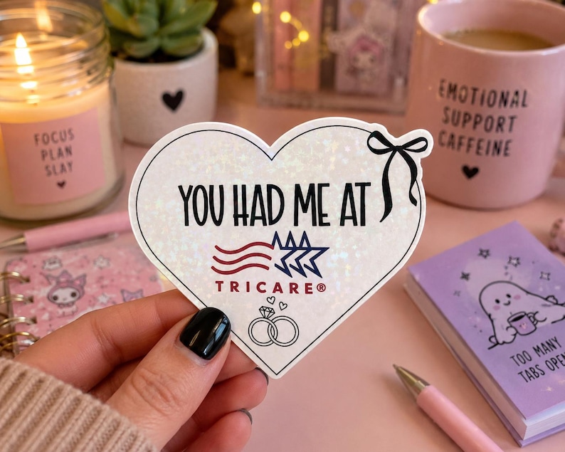 May include: A heart-shaped sticker with a holographic finish featuring a black bow, the text "YOU HAD ME AT", a red and blue TRICARE logo, and an illustration of two interlocking rings with two small hearts above them on a white background.