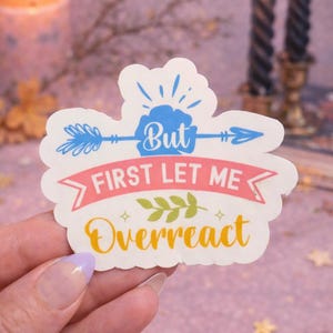 May include: A white sticker with the text "But First Let Me Overreact." The design includes a blue arrow, a pink banner, and green leaves. The sticker is held in a hand with a lavender-tipped nail.