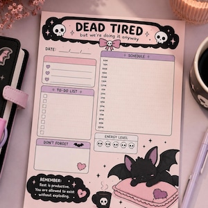 May include: A rectangular daily planner notepad with a light pink background. The header reads DEAD TIRED but we're doing it anyway with skull icons. It includes sections for date, schedule from 6AM to 10PM, a to-do list, energy level, and a don't forget box.