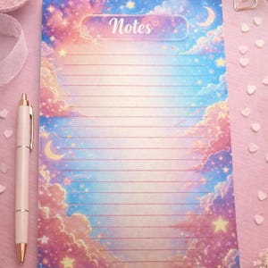 May include: Lined notepad with a dreamy sky design. The notepad features a pastel color palette of pink, blue, and yellow, with clouds, stars, and a crescent moon. The word "Notes" is written at the top. A white pen is next to the notepad.
