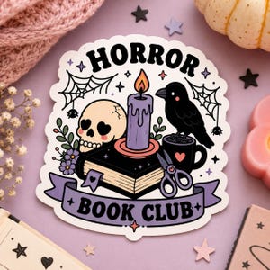 May include: A die-cut sticker featuring the text HORROR BOOK CLUB. The illustration includes a beige skull with heart-shaped eyes, a purple candle, a black book, a black crow perched on a mug, spiderwebs, and scissors on a light purple background.