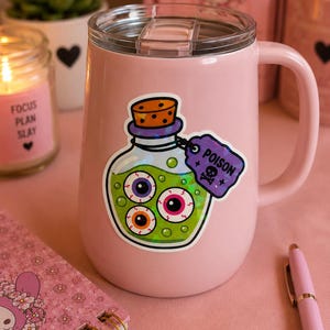 May include: A light pink insulated tumbler with a handle and a clear lid. A holographic sticker on the front depicts a glass bottle filled with green liquid and three eyeballs, featuring a purple tag labeled POISON with a skull and crossbones.