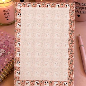 May include: A rectangular notepad featuring a central area with a faint ghost and floral pattern, surrounded by a border of white ghosts wearing flower crowns and holding mugs, interspersed with small orange and pink flowers on a light pink background.