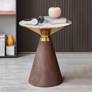 May include: A modern side table with a round marble top and a gold-colored trim. The table has a conical wooden base and is topped with a decorative apple, a book, and headphones. The table is a stylish piece of furniture.