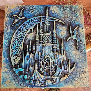 May include: A square, textured artwork featuring a detailed, raised relief of a fantasy castle. The castle is set within a crescent moon shape, with two stylized birds. The color palette includes blues, gold, and hints of white.