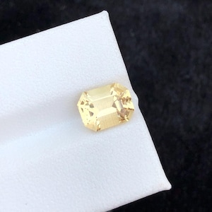 May include: A rectangular, pale yellow gemstone with an emerald cut. The gem is set against a white surface, with a dark background. The gemstone has a brilliant, faceted appearance, reflecting light.