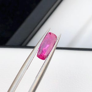 Peut inclure: Un rubis de forme rectangulaire, maintenu par une pince. La pierre précieuse affiche une couleur rose intense et présente des facettes qui captent la lumière. Le fond est un mélange de blanc et de noir, créant un contraste.