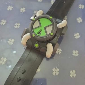 May include: A black and green toy watch with a circular face featuring a green and gray design. The watch has a black strap and white accents. The watch is a toy.