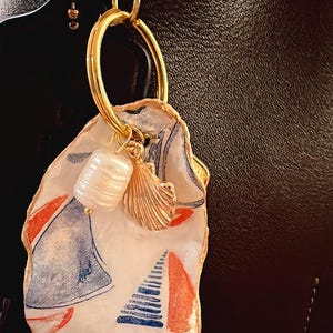 May include: A unique keychain featuring a sailboat design on a white background. The keychain has a gold-colored clasp and ring, with a pearl and seashell charm. The sailboat design includes red and blue accents.