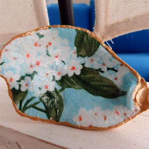 May include: A decorative oyster shell dish with a floral design. The interior features a light blue background with white flowers and green leaves. The shell's edge is painted gold, creating a unique and artistic piece.