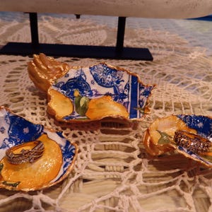 Op de afbeelding: Drie decoratieve oesterschelpen ringhouders. Elke schelp is beschilderd met een blauw en wit bloemmotief, oranje fruit en gouden accenten. Eén schaal bevat een gouden ring. De schelpen liggen op een kanten oppervlak.