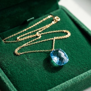 Op de afbeelding: Een gouden ketting met een enkele, gefacetteerde, kussenvormige lichtblauwe edelsteen als hanger. De ketting ligt in een met donkergroen fluweel bekleed doosje, wat het contrast tussen het goudkleurige metaal, de blauwe steen en de stof benadrukt.