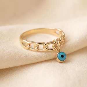 Puede incluir: Un anillo de tono dorado con un diseño de banda de eslabones de cadena. Un pequeño dije circular cuelga del centro de la banda, mostrando un motivo de ojo azul y blanco con una pupila negra. El metal tiene un acabado pulido y brillante.