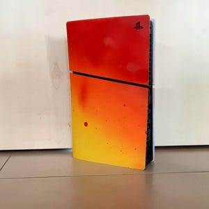 Może przedstawiać: Konsola PlayStation 4 z żywym gradientem. Schemat kolorów przechodzi od czerwonego u góry do żółtego u dołu. Konsola stoi pionowo na tle białej ściany i kafelkowej podłogi.