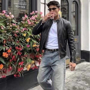 May include: A person wearing a black leather jacket, white polo shirt, and light blue jeans. The person is holding a cup and standing in front of a building with a floral display. The building has a sign that says "SUNDAYS WE ROAST".