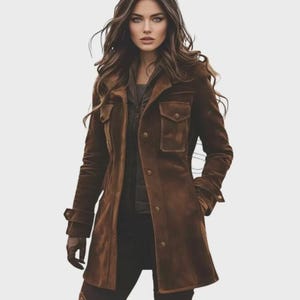 May include: A brown suede coat with a button-up front, two chest pockets, and long sleeves. The coat is worn over a dark top and black pants. The model is wearing gloves. The coat has a classic, tailored design.