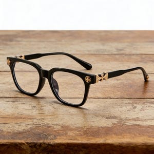 May include: Black-framed spectacles with gold-coloured cross detailing on the temples. The glasses have a square shape and are displayed on a wooden surface.