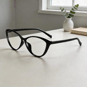 May include: Black cat-eye glasses with clear lenses. The frames have a modern design and are set on a white surface. The glasses are positioned in front of a window and a small vase with greenery. The overall aesthetic is minimalist.