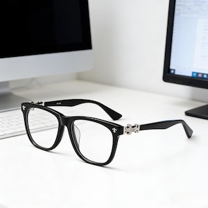 Op de afbeelding: Zwarte bril met vierkante montuur en zilveren details op de pootjes. De glazen zijn helder en de bril staat tegen een witte achtergrond. Het ontwerp bevat een decoratief element op de pootjes.