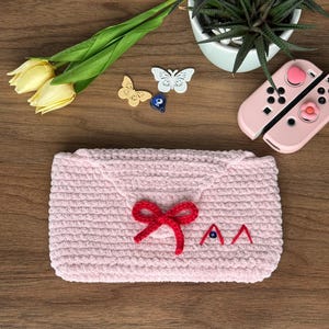 Könnte beinhalten: Eine hellrosa, gehäkelte Hülle im Umschlagstil mit einer roten Schleife und gestickten Details. Die Hülle ist rechteckig und hat einen Klappenverschluss. Das Bild enthält auch ein Paar rosa Gamecontroller, gelbe Tulpen und dekorative Schmetterlinge.