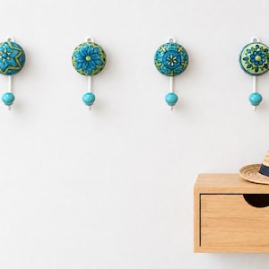 Op de afbeelding: Vier wandhaken met ronde keramische knoppen in blauwe en groene bloemen- en geometrische patronen. Elke haak heeft een witte metalen steel en een kleine turquoise kogelvormige punt. Een strooien hoed ligt op een ladekast van licht hout.