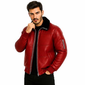 Puede incluir: Una chaqueta bomber de cuero rojo con cuello de piel sintética negra, cremallera frontal plateada y un bolsillo con cremallera en la manga izquierda. La chaqueta se lleva sobre una camiseta negra, con pantalones negros y botas de cuero negro.