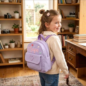 May include: A lilac corduroy backpack with the name "Maigie" embroidered on the front, featuring floral and heart embellishments. The backpack has a front pocket and purple straps. The child is wearing a cream sweater and blue jeans.