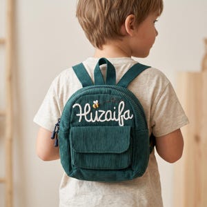 Könnte beinhalten: Ein dunkelgrüner Rucksack aus Kord mit dem gestickten Namen Huzaifa in weißer Schreibschrift und einem kleinen Bienenmotiv. Die Tasche verfügt über eine Vordertasche mit Klappe, einen Tragegriff und verstellbare Schultergurte mit dunkelblauen Reißverschlussanhängern.