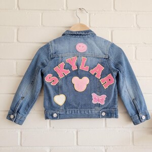 Peut inclure: Une veste en jean bleu avec le nom SKYLAR écrit en lettres roses de style universitaire au dos. La veste est ornée de plusieurs écussons, dont un visage souriant rose, une silhouette de tête de souris rose, un cœur blanc et un papillon rose.