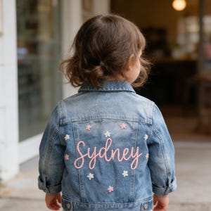 Op de afbeelding: Lichtblauwe spijkerjas met de naam "Sydney" in roze script op de rug geborduurd. De jas is versierd met kleine witte en roze bloemenapplicaties. De kinderjas heeft een klassieke kraag en manchetten met knopen.