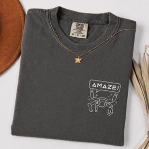 May include: A dark gray Comfort Colors t-shirt with a gold star pendant necklace. The shirt features a white graphic of a robot with the word "AMAZE!" in a speech bubble. The shirt is folded.
