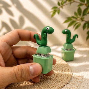 May include: Green dinosaur-shaped keycap on a green square base. The keycap has a cartoonish design with a friendly expression. The image shows two keycaps, one held by a hand and another in the background. The keycaps are likely for a mechanical keyboard.