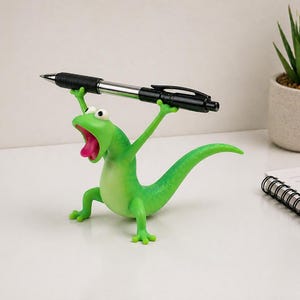 Puede incluir: Una caprichosa figura de lagarto verde con la boca muy abierta y los brazos extendidos, sosteniendo un bolígrafo negro. El lagarto tiene una lengua rosa y ojos blancos. El bolígrafo es negro y plateado. Un divertido accesorio de escritorio.