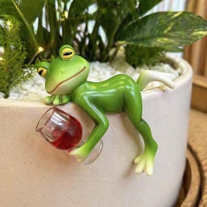 Puede incluir: Una caprichosa figura de rana verde con ojos amarillos, sosteniendo un pequeño vaso de líquido rojo. La rana está posada en el borde de una maceta de color claro, añadiendo un toque de diversión a cualquier exhibición de plantas.