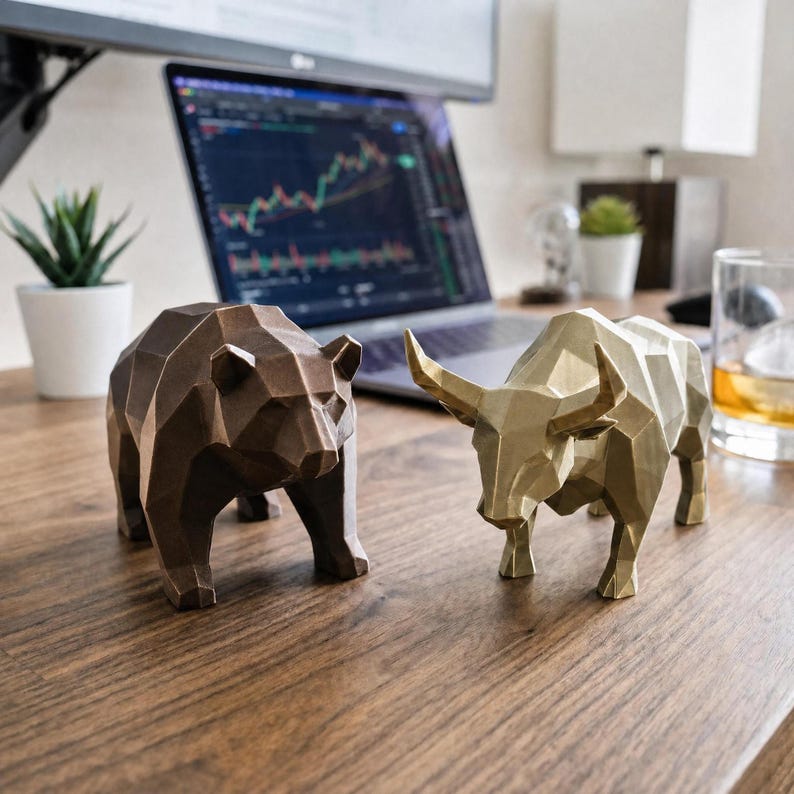 A bronze geometric bear and a gold geometric bull statue are on a wooden desk, with a laptop displaying stock charts and a glass of whiskey in the background.