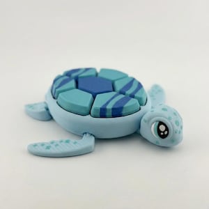 Peut inclure: Figurine de tortue bleu clair avec une carapace segmentée dans les tons de bleu. La carapace présente un motif géométrique avec des rayures bleu foncé. La tortue a un visage détaillé avec de grands yeux et des taches.