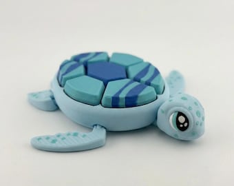 Puede incluir: Figura de tortuga azul claro con caparazón segmentado en tonos azules. El caparazón tiene un diseño geométrico con rayas azul oscuro. La tortuga tiene una cara detallada con ojos grandes y manchas.