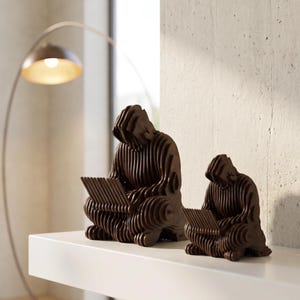 May include: Two brown wooden sculptures of a person reading, with a layered design. The sculptures are on a white shelf, with a modern arc lamp in the background. The figures are in a seated position, focused on their books.