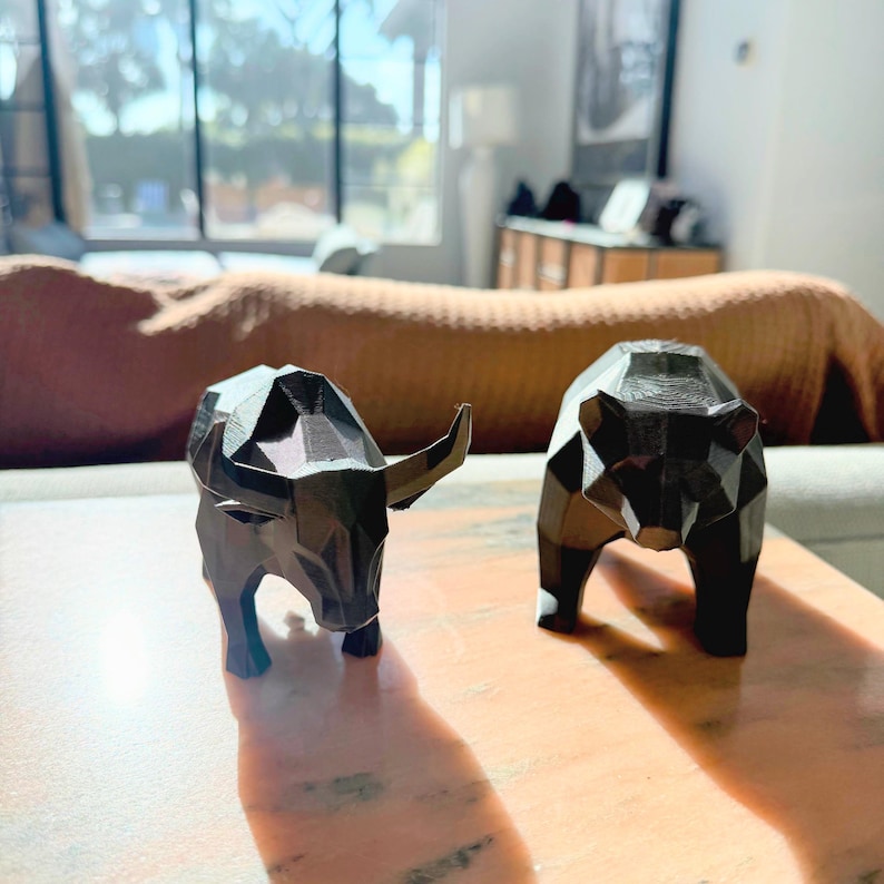 A black faceted bull and bear statue sit on a light brown table, with a window and couch in the background.