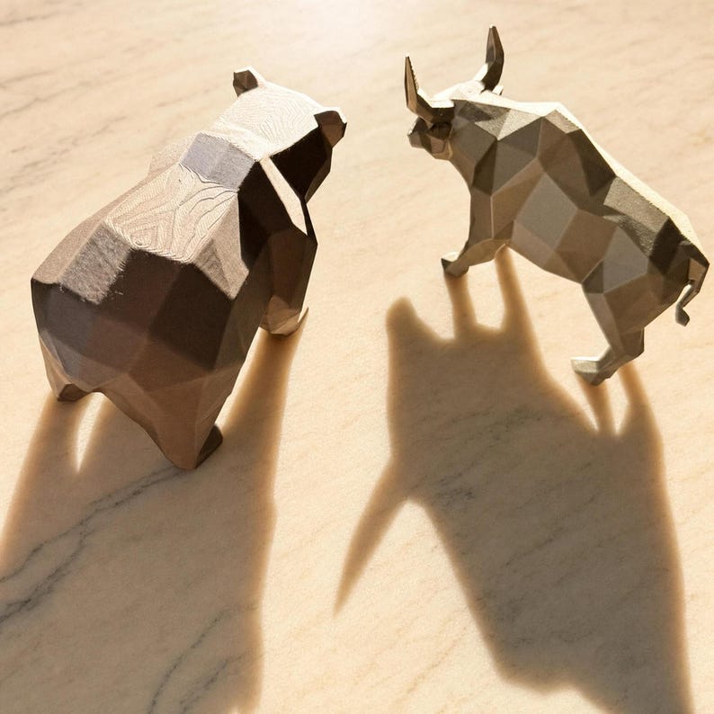 A bronze geometric bear and a light gray geometric bull statue face each other on a light marble surface, casting long shadows.