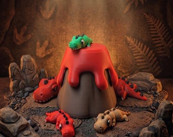 Puede incluir: Un diorama con un volcán con lava roja y varias pequeñas figuras de dinosaurios. Los dinosaurios son rojos, verdes y marrones, y están colocados alrededor del volcán sobre una superficie rocosa. El fondo es una cueva.